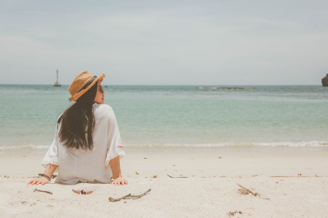 Žena v bielej blúzke a klobúku sedí na pláži pri mori.jpg
